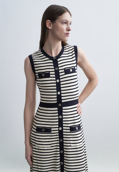 Jeune femme portant une robe sans manches à rayures horizontales noires et blanches, avec des boutons et des poches, debout, une main sur la hanche, regardant vers la droite.