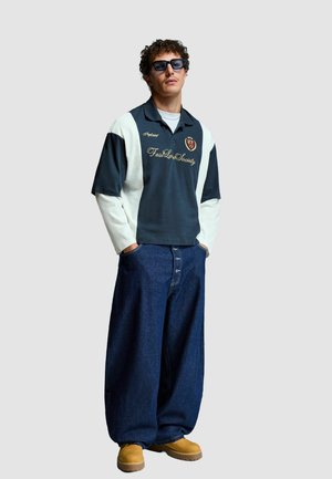 Zwart-wit polo met geborduurd logo, gecombineerd met loszittende blauwe spijkerbroek en tan schoenen. Accessoires omvatten donkere zonnebril.