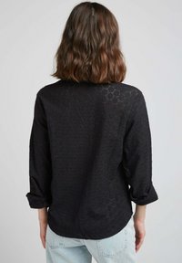 Blouse noire à manches longues avec un motif texturé en mailles ouvertes. Présente des manches retroussées et un ourlet arrondi. Coupe décontractée avec une silhouette relaxée.