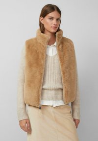 Jonge vrouw in een tan nepbont vest over een crèmekleurige gebreide trui en een wit shirt, gecombineerd met een beige corduroy rok tegen een effen achtergrond.