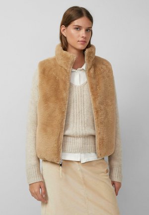 Jonge vrouw in een tan nepbont vest over een crèmekleurige gebreide trui en een wit shirt, gecombineerd met een beige corduroy rok tegen een effen achtergrond.