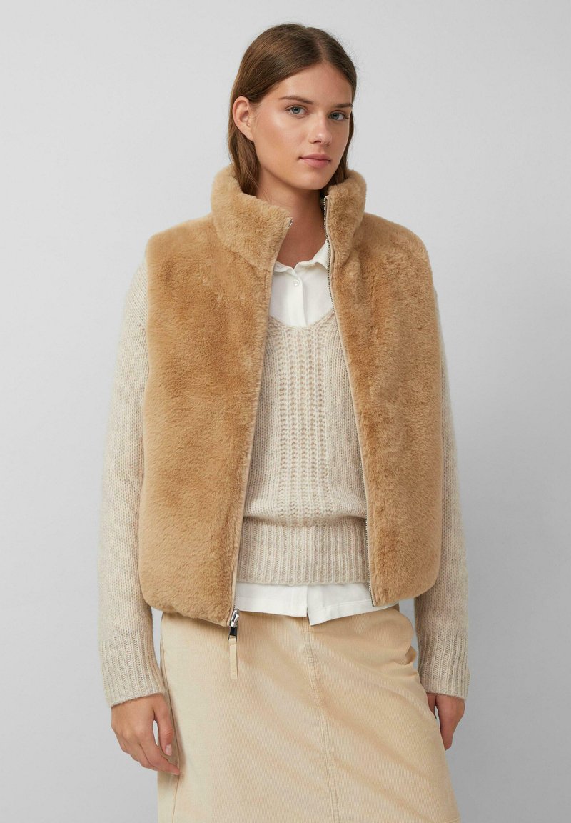 Jonge vrouw in een tan nepbont vest over een crèmekleurige gebreide trui en een wit shirt, gecombineerd met een beige corduroy rok tegen een effen achtergrond.