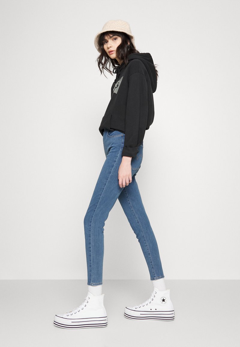 Schwarzer Hoodie mit grafischen Details, blaue Skinny-Jeans und weiße High-Top-Sneaker mit schwarzen Streifen. Modell posiert im Profil mit einem beigen Hut.