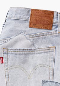 Gai zila denima bikses ar brūnu ādas uzlīmi, kurā redzams Levi's logs. Izteiksmīga arku šuvju raksts aizmugurējās kabatās. Pievienota sarkana узlīme.