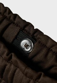 Brūns audums ar viļņotu maliņu, ar caurspīdīgu četrcauruļu pogu un melnu elastīgu joslu. Strukturēta virsma ar šuvju detaļām.