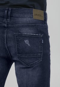 Mörkblå denimjeans har en slitna ficka, en brun varumärkeslapp vid midjan och en subtil, använd textur.
