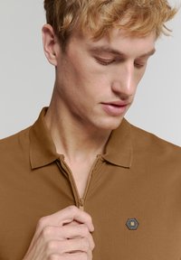 Bruine polo met ritssluiting van een textuurstof, met een kraag en een hexagonaal logo-accent nabij de rits. Handen houden de kraag vast.