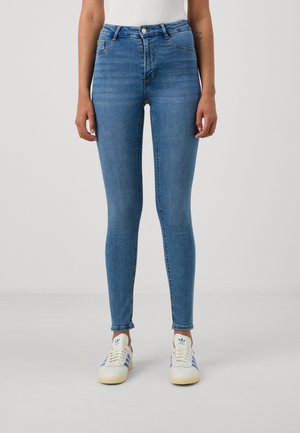 Žena oblečená v úzkých světlomodrých džínách a bílých teniskách s modrými pruhy, stojící před jednobarevným světlým pozadím.