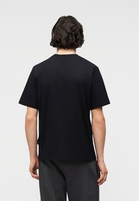 Άνδρας με μαλλιά μέχρι τους ώμους, φοράει απλό μαύρο κοντομάνικο μπλουζάκι και σκούρο παντελόνι, από πίσω σε απλό φόντο.