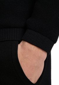 Tessuto di maglione nero a maglia, con polsini a coste e dettaglio della tasca. La trama è liscia con un motivo uniforme in tutto il capo.