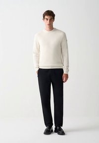 Crèmekleurige gebreide trui met geribbelde manchetten en zoom, gecombineerd met zwarte maatbroek en zwarte schoenen. Eenvoudig, modern ontwerp en neutrale kleuren.