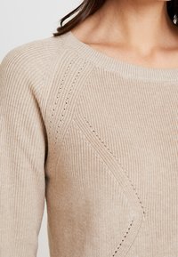 Kobieta w beżowym swetrze z dzianiny z wzorzystymi detalami wokół ramienia i klatki piersiowej, odsłaniająca szyję i górną część ramienia.