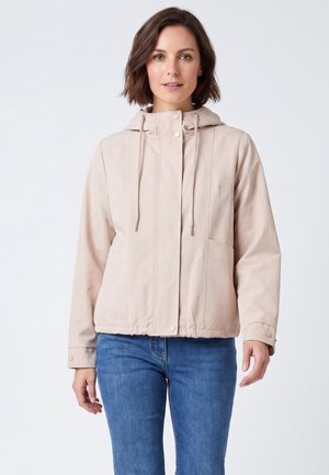 Donna con giacca beige con cappuccio e bottoni automatici e jeans blu, in piedi davanti a uno sfondo bianco uniforme.
