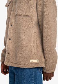 Tan fleece jacket with a textured finish, featuring a chest pocket and branded metal tag, paired with blue denim jeans.