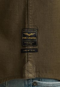 Tkanina w oliwkowym kolorze o tkanym wzorze z czarną metką, na której złotymi literami widnieje napis "PME LEGEND" oraz "AMERICAN CLASSIC", z detalami stembowania.