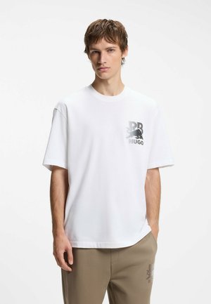 Jonge man met kort haar, die een wit T-shirt draagt met een klein zwart logo op de borst en beige broek, staand tegen een effen achtergrond.