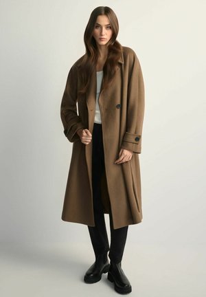 Brązowy wełniany płaszcz trencz o luźnym kroju, z podwójnym zapięciem na guziki i klapami z wycięciem, noszony na czarnych spodniach i masywnych czarnych butach.