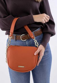 Bruine leren schoudertas met een ronde vorm, gouden hardware en een gestructureerd oppervlak, vastgehouden door een persoon in een donkere trui en blauwe jeans.