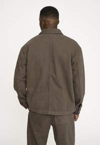 Brun bomuldsjakke med en afslappet pasform, synlige sømme og en krave. Bagsiden har en yoke-design og sidelommer. Flad tekstur.