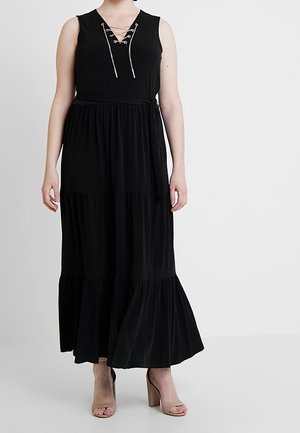 Femme portant une robe maxi noire sans manches à volants avec un col lacé et des sandales nude à talons blocs, debout sur un fond blanc.