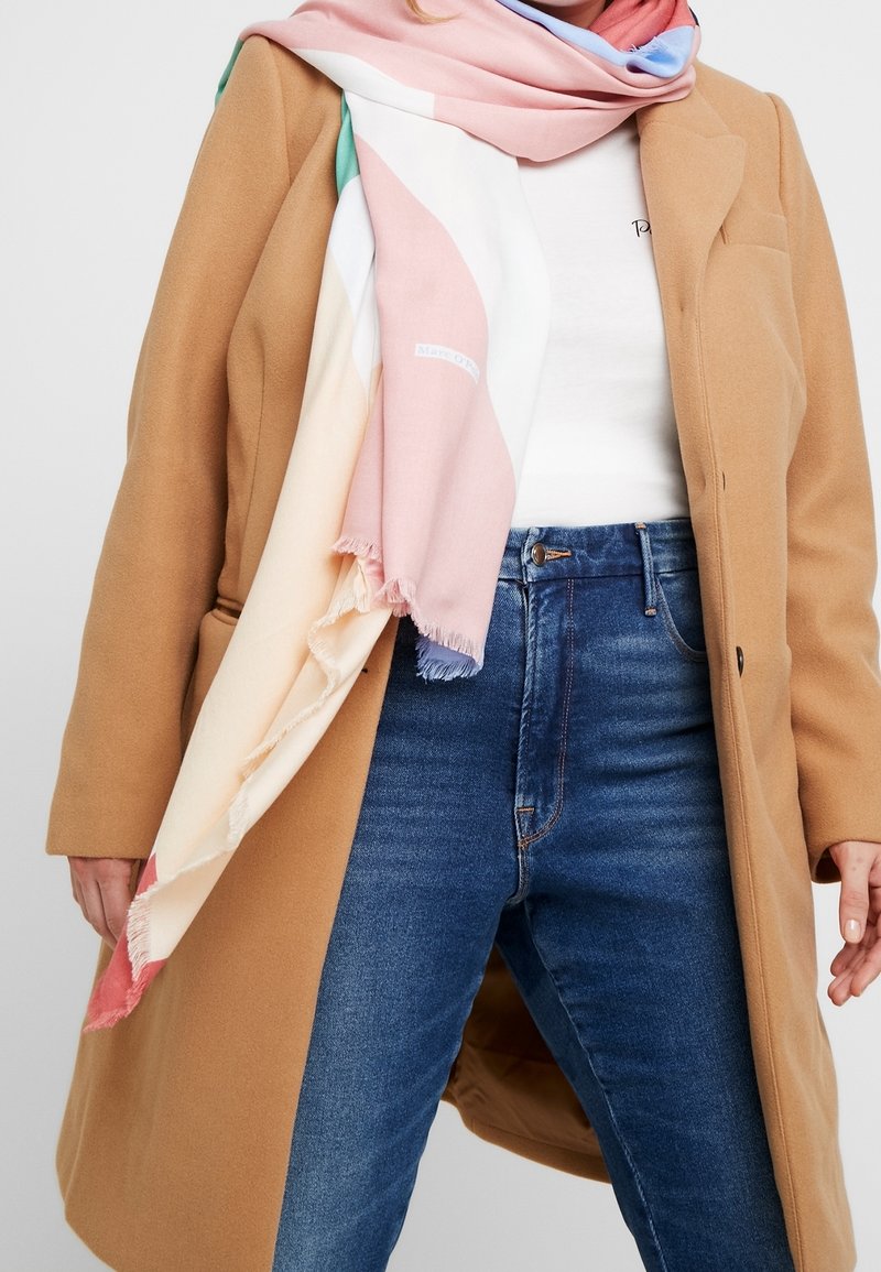 Femme portant un manteau camel, un jean bleu, un haut blanc, et une écharpe aux couleurs pastel enroulée autour du cou, debout les mains détendues.