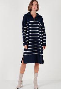 Rochie tricotată în dungi albastre și albe, cu guler cu fermoar, mâneci lungi, slituri laterale și o croială lejeră, asortată cu cizme glossy bej până la gleznă.
