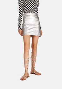 Nainen, jolla on päällä hopeinen minihelma, musta-valkoinen ruutukuosisella pitkähihaisella paidalla ja metalliväriset nauhakenkämatalat sandaalit, seisoo valkoista taustaa vasten.