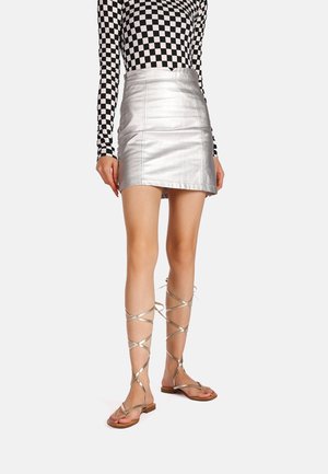 Vrouw in een zilveren minirok, zwart-witte geblokte lange mouw top en metallic vetersandalen met platte zool, staande tegen een witte achtergrond.