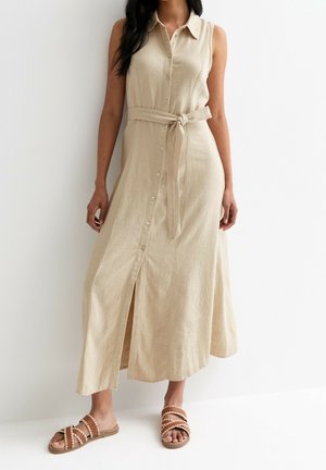 Ærmeløs beige midi-kjole med knapper og bundet stofbælte, båret med brune flade sandaler med flettede stropper.