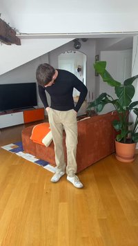 Άνδρας με μαύρο μακρυμάνικο πουκάμισο και μπεζ παντελόνι, στέκεται σε σαλόνι με πορτοκαλί καναπέ, πράσινο φυτό, τηλεόραση και ξύλινο πάτωμα.