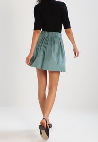 Světle zelená sukně se sklady a elastickým pasem, vyrobená z hladké tkaniny. V kombinaci s přiléhavým černým topem a černými sandály.