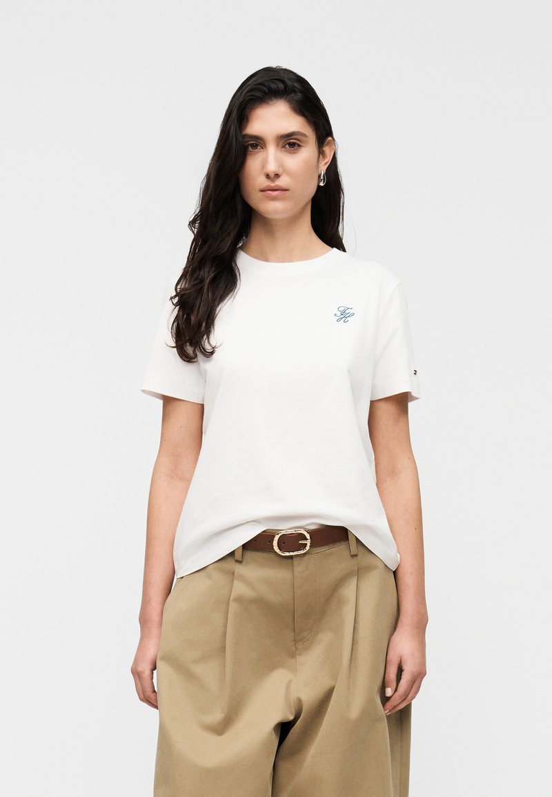 Jeune femme aux longs cheveux foncés portant un t-shirt blanc à manches courtes avec un logo bleu et un pantalon beige taille haute avec une ceinture marron.