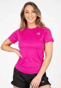 Korte mouwen sportshirt van helderroze stof, met een grijs logo op de bovenste linkerzijde en een gladde, ademende textuur. Draagt het met zwarte shorts.