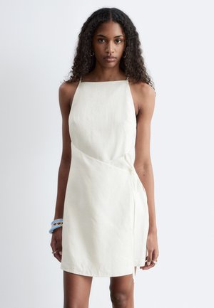 Vrouw met krullend haar, die een mouwloze, witte, knielange jurk draagt, staande tegen een effen lichtgrijze achtergrond.