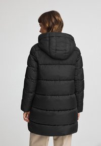Czarna kurtka puchowa z kapturem, o pikowanym wzorze, wydłużonym kroju i gładkiej fakturze. Wyraźna dzięki grubej, izolowanej wyściółce.