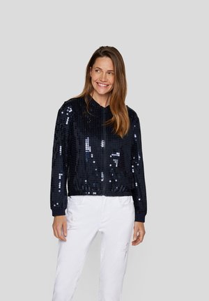 Marineblaue Pailletten-Bomberjacke mit Reißverschluss vorne, gerippten Manschetten und Saum. Präsentiert eine glänzende Textur mit unterschiedlichen Lichtreflexionen. Kombiniert mit weißen Hosen.