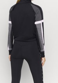 Chaqueta deportiva negra con detalles en rosa; presenta patrones geométricos en las mangas; hecha de una tela suave y elástica; diseño de cuello alto.