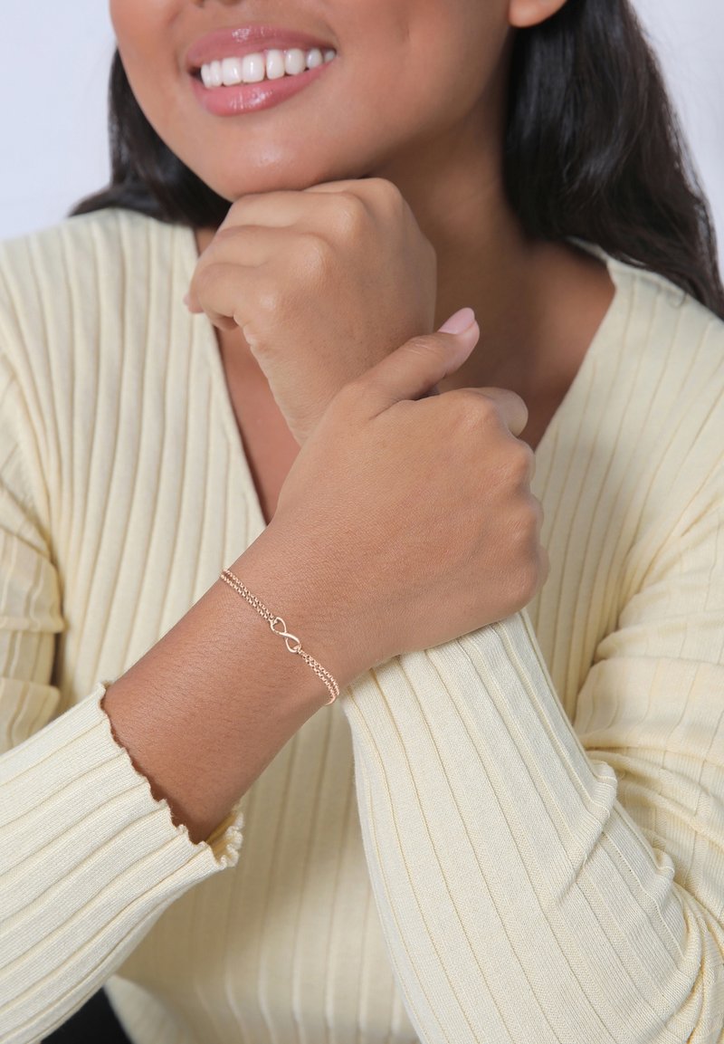 Femme portant un haut côtelé à manches longues de couleur jaune clair sourit, mettant en valeur un délicat bracelet infini en or à son poignet.