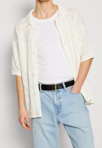 Witte gebreide knopencardigan met een ruitpatroon, korte mouwen, gedragen over een wit T-shirt, gecombineerd met lichtblauwe jeans en een zwarte riem.