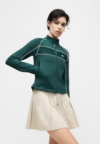 Γυναίκα που φοράει πράσινο ζακέτα με φερμουάρ και λευκή επένδυση, και μπεζ πλισέ φούστα, ποζάροντας με το ένα χέρι στην τσέπη της ζακέτας μπροστά σε απλό φόντο.