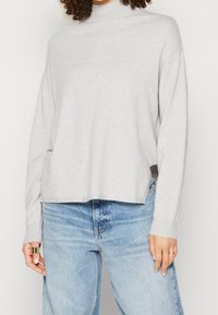 Ljusgrå pullovertröja, mock neck, mjukt stickat material, avslappnad passform, sidofickor vid fållen, bärs med ljusblå denimjeans.