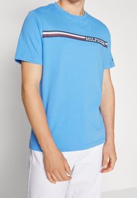 Blå kortärmad t-shirt i bomull. Har en horisontell rand med röda och marinblå accenter, som visar "HILFIGER"-logotypen.