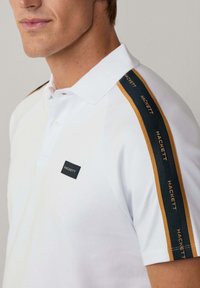 Hvid polo t-shirt med et sort logo på brystet og kontrasterende grønne og guld striber langs skuldersømmene. Blødt bomuldsstof.