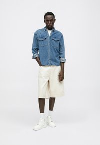 Denimhemd mit Knopfleiste, zwei Brusttaschen; kombiniert mit cremefarbenen Shorts und weißen Sneakern. Einfaches Design, lockere Passform, minimaler Textur.