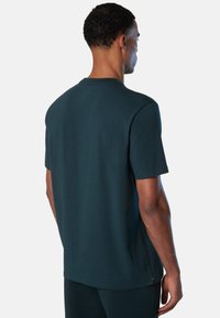 Hombre con pelo corto y rizado, vistiendo una camiseta de manga corta de color verde azulado oscuro y pantalones a juego, de pie de espaldas sobre un fondo claro y sencillo.