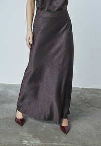Maxiskirt i mørkebrun satin med et tekstureret mønster. Har en høj talje og et flydende design, der er parret med spidse burgunderhæle.