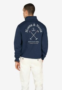 Pulóver de forro polar azul marino con cuello alto, que presenta texto y gráficos en blanco de un sombrero y palos de golf cruzados en la parte posterior. Pantalones blancos visibles.
