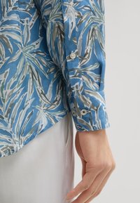 Kortebloem blauw shirt met een bladerenpatroon in wit en grijs. Knopmanchetten en een zachte textuur. De zoom van het shirt is licht gebogen aan de onderkant.