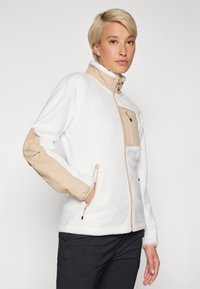 Giacca in felpa bianca con accenti in nylon beige sulle maniche e sulla tasca del petto, dotata di un colletto alto e chiusure con zip.