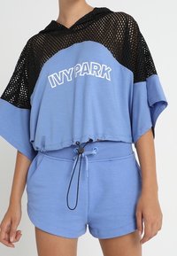 Blå Ivy Park-shorts och matchande kort topp med svart mesh-hood, bärs av en person med avslappnade händer vid sidorna.